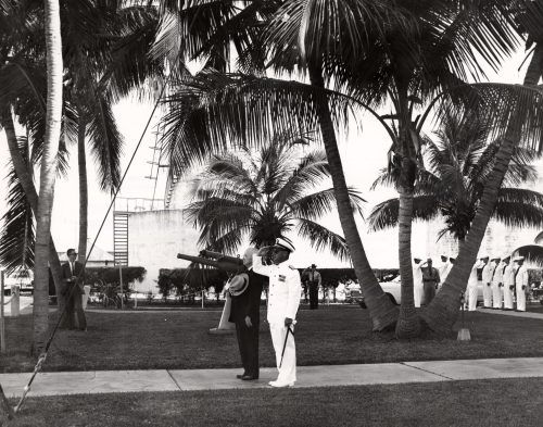 A man in a white dress Navy uniform salutes standing next to another man in a dark suit holding his hat, with palm trees, guns and sailors in the background.