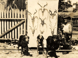 A man and dogs sitting outside a house with deer skulls mounted on a board.