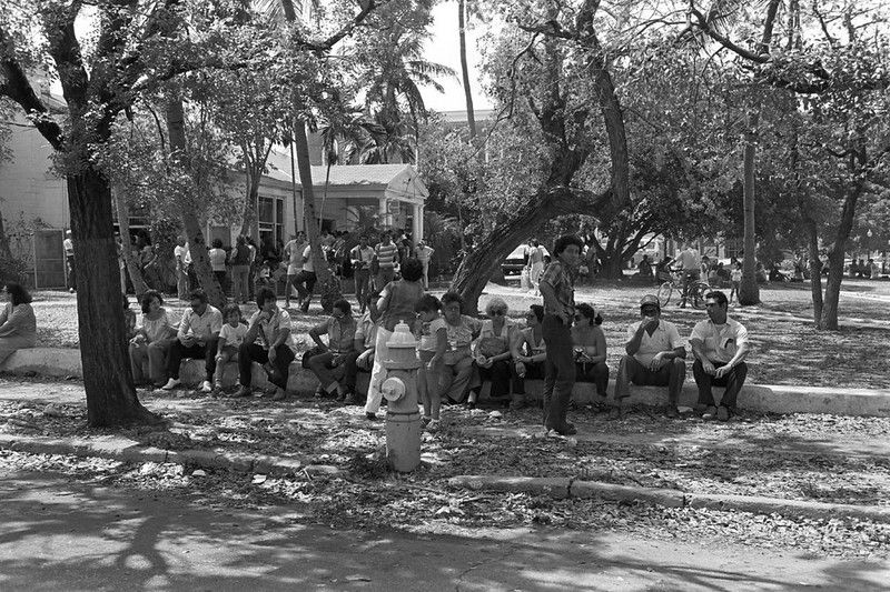 A line of people sitting on a low wall in front of a building with a group of people standing in front of it.