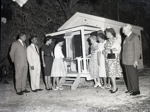 A group of people watch a woman cut the ribbon on the porch of a playhouse