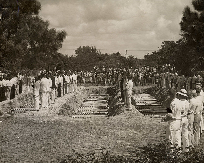 A large group of people at a mass burial with three rows of caskets being interred.