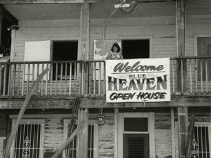 A woman standing on the upstairs porch of a house with a sign that reads Welcome Blue Heaven Open House