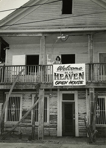 A woman on the upstairs porch of a building with a sign that reads Welcome Blue Heaven Open House.
