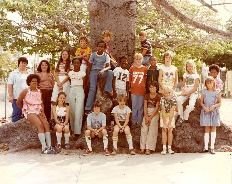 A group of kids sitting at the roots of a big tree.
