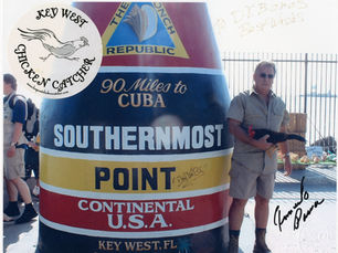 A man holding a chicken next to the southernmost point monument, with a seal that reads Key West chicken catcher.