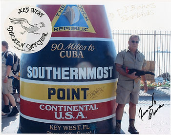 A man holding a chicken next to the southernmost point monument, with a seal that reads Key West chicken catcher.