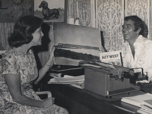 A woman sitting in front of a desk and a man sitting behind the desk smile at each other.