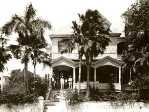 People standing on the porch of a two story house with palm trees in front of it.