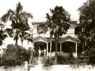 People standing on the porch of a two story house with palm trees in front of it.
