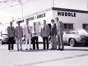 Seven men lined up outside a building with lettering on the sides reading Huddle Pontiac Buick.