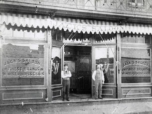 Two men stand in front of a storefront with Jack's Saloon, Finest Brands Wines, Liquors & Cigars written in the windows.