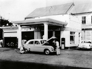 Two men with a mid 20th century style car with its hood up in front of a service station.