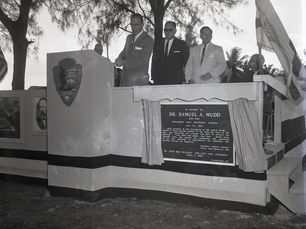 Three men at a podium with a National park Service shield and a plaque in memory of Dr. Samuel A Mudd.