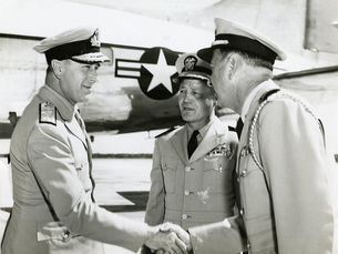 Two men in uniform shake hands in front of a third man in uniform