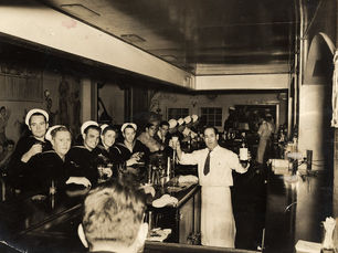 A bar lined with men mostly in Navy uniforms, while a bartender behind the bar holds up a bottle.
