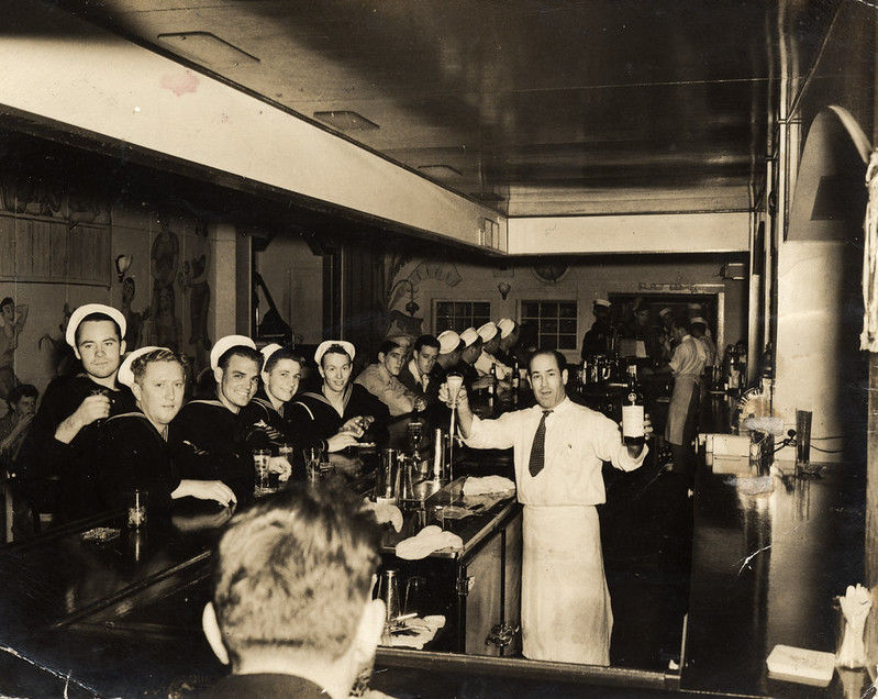 A bar lined with men mostly in Navy uniforms, while a bartender holds up a bottle.