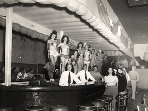 A bar with women in bikins standing on the bar, bartenders behind the bar and patrons on stools.
