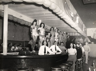 A bar with women in bikins standing on the bar, bartenders behind the bar and patrons on stools.