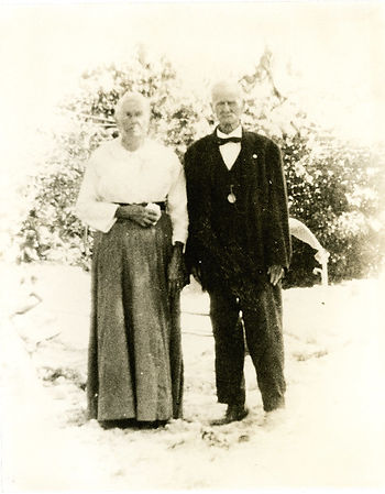 An older woman in a long skirt and an older man in a suit and bow tie stand outside.