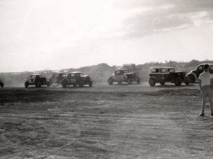 A person stands in the foreground in front of cars with numbers on a dirt track.