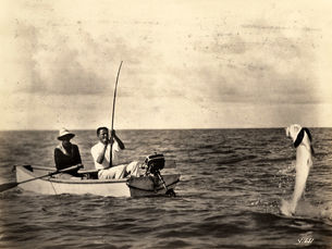 Two people in a small boat. One has a fishing rod with a large fish on the line.