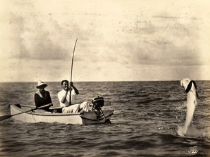 Two people in a small boat. One has a fishing rod with a large fish on the line.
