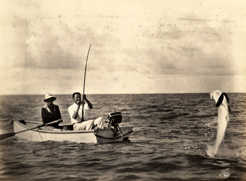 Two people in a small boat. One has a fishing rod with a fish on the line.