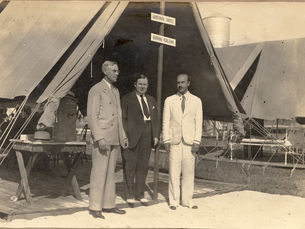 Three men in suits standi n front of a tent on a platform.