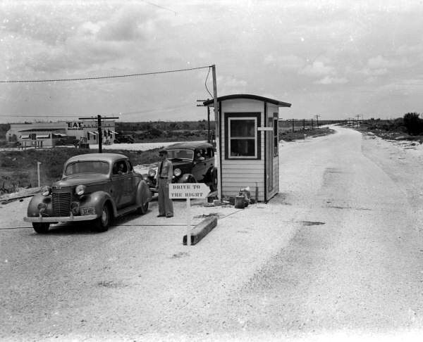 Two 1930s style cars and a person in a uniform at a tollboth with a sign that reads drive to the right.