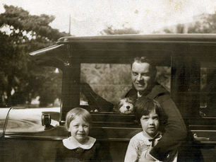 A man in a car with a dog on his lap and two children in front of the car