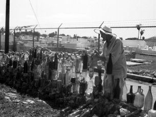 A wpman stands in front of a fence made of bottles with a chainlink fence and cemetery behind her.