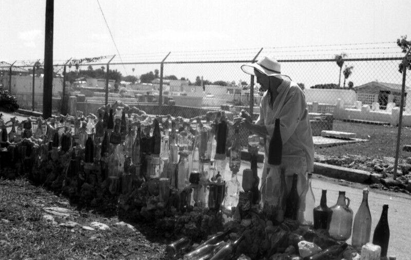 A wpman stands in front of a fence made of bottles with a chainlink fence and cemetery behind her.