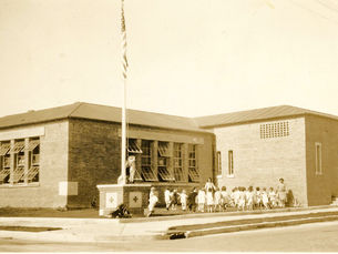 A brick building with a line of children, two adults, and a flagpole in the yard.