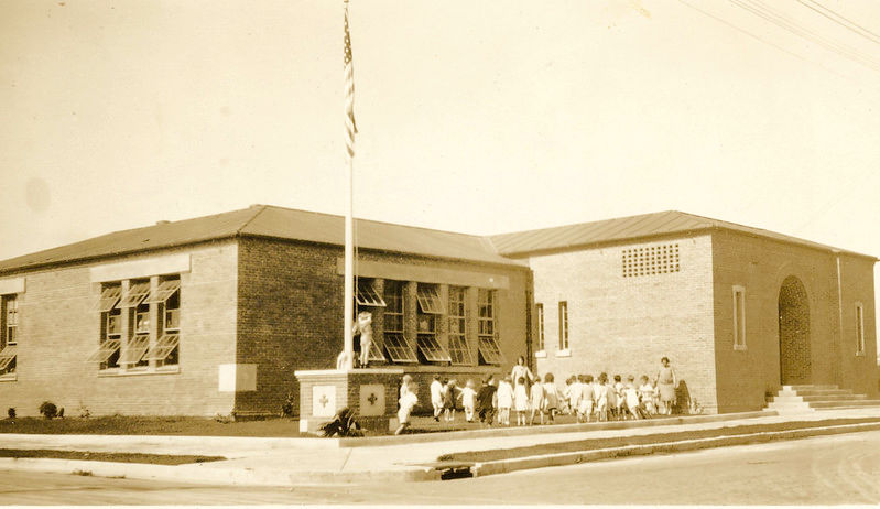 A brick building with a line of children, two adults and a flagpole in the yard.