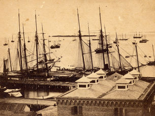 Aerial view of building roofs on a shoreline and sailing ships in a harbor.
