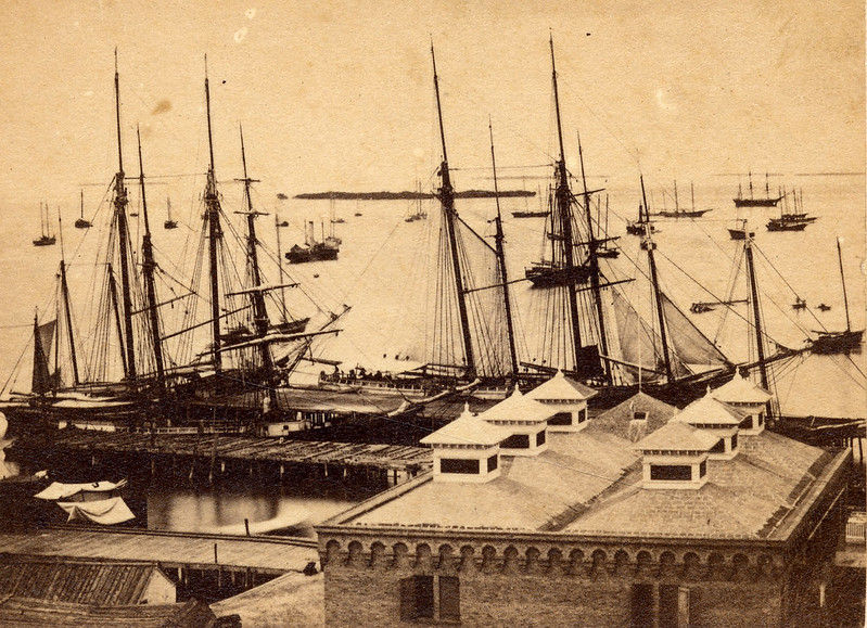 Aerial view of roofs on a shoreline and sailing ships in a harbor.