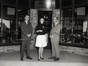 Three people in front of a storefront while the woman in the middle cuts a ribbon.
