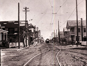 An unpaved street with buildings on the side and tracks and an electric trolley 