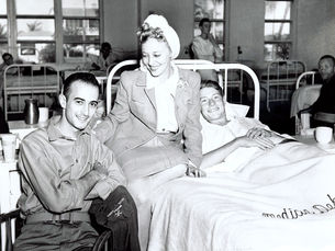 A young woman sits on a hospital bed with a young man in the bed and another sitting in a chair in front of her.