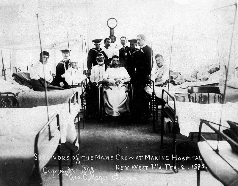 A group of men, some in Navy uniforms, in a hospital ward. Text on photo reads Survivors of the Maine Crew at Marine Hospital, Key West Fla. Feb. 21. 1898. Copyright 1898 Geo. C. Mages, Chicago.