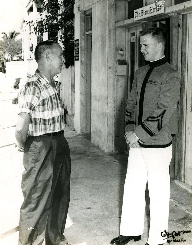 Two men, one in uniform, in front of a building with a sign that reads Miami Herald.