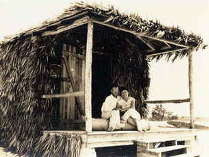A couple sitting on the porch of a cabana covered in palm fronds.