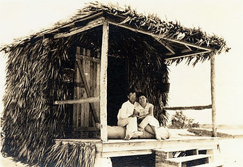 A couple sitting on the porch of a cabana covered in palm fronds.