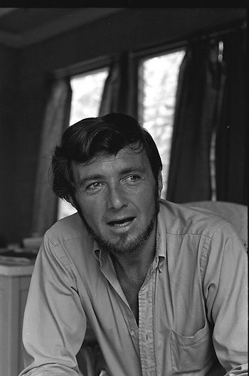 Black-and-white portrait of a man leaning forward indoors, with curtained windows behind him.