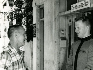 Two men, one in uniform, in front of a building with a sign that reads Miami Herald
