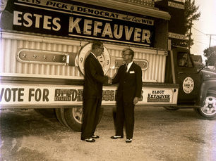 Two men shake hands in front of a truck with a sign that says Estes Kefauver