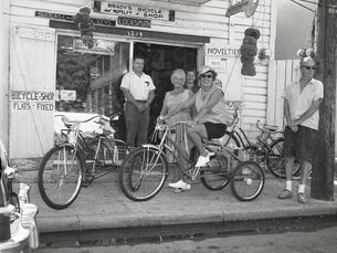 A group of people with tricycles in front of a shop with a sign that reads Brady's Bicycle and Novelty shop.