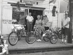 A group of people with tricycles in front of a shop with a sign that reads Brady's Bicycle and Novelty shop.