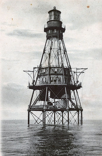 Black-and-white photo of a tall offshore lighthouse on steel stilts rising from calm sea under a cloudy sky.