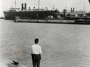 A person standing at a dock looking across the water at a steamship at a dock.